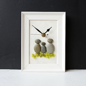 May include: A white framed clock featuring a family of three, represented by three grey stones. The stones are arranged to resemble a family with a child in the centre. The clock is set against a black background.