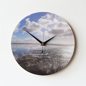 May include: A round clock with a black metal frame and hands. The clock face features a photograph of a cloudy sky reflected in a body of water.