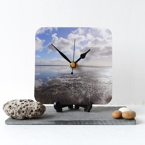 May include: A square clock with a black face and gold hands. The clock face features a photograph of a beach with a cloudy sky and a reflection of the clouds in the water.