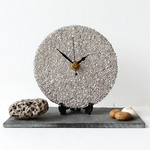 May include: A circular clock with a silver-toned face and black hands, set on a black stand. The clock's face is covered in small, reflective pieces. The base is a grey rectangular slate, with decorative stones.