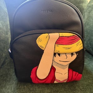 May include: A black backpack with the brand name TELENA embossed on the top front. The front pocket features a hand-painted illustration of a character wearing a yellow straw hat with a red band and a red shirt, with one arm raised to the hat.