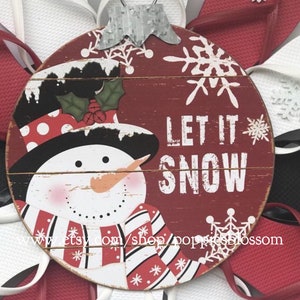 May include: A round, red wooden ornament with a snowman design and the words "LET IT SNOW". The snowman has a black hat, red and white scarf, and an orange carrot nose. The ornament is surrounded by red and white flip-flops.