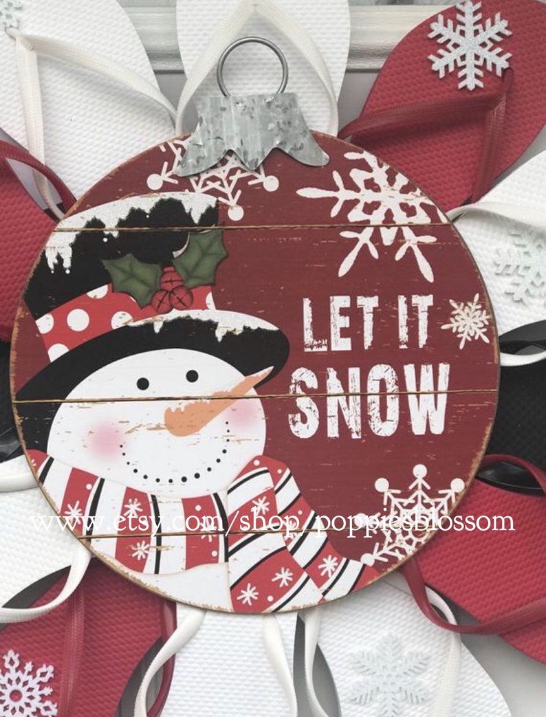 May include: A round, red wooden ornament with a snowman design and the words "LET IT SNOW". The snowman has a black hat, red and white scarf, and an orange carrot nose. The ornament is surrounded by red and white flip-flops.