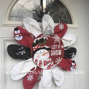 May include: A festive Christmas wreath made of red, white, and black flip-flops arranged in a circular pattern. A central ornament features a snowman and the words "Let it Snow".