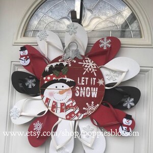 May include: A festive Christmas wreath made from red, white, and black flip-flops, arranged in a flower shape. The wreath features a central wooden ornament with a snowman and the words "Let It Snow." Snowflake accents and small snowman embellishments complete the holiday design.