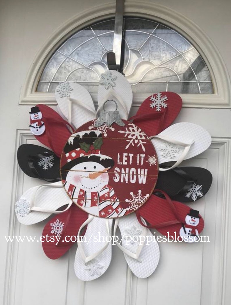 May include: A festive Christmas wreath made from red, white, and black flip-flops, arranged in a flower shape. The wreath features a central wooden ornament with a snowman and the words "Let It Snow." Snowflake accents and small snowman embellishments complete the holiday design.