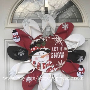 May include: A festive Christmas wreath made of red, white, and black flip-flops arranged in a circular pattern. The center features a wooden ornament with a snowman design and the words "LET IT SNOW". Decorative snowflakes and snowman accents complete the holiday theme.