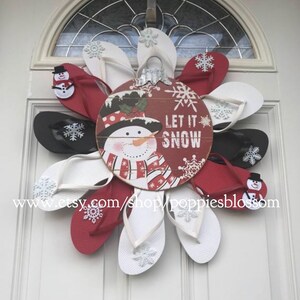 May include: A festive winter wreath made of red, white, and black flip-flops arranged in a circular pattern. A wooden ornament in the center features a snowman and the words "LET IT SNOW". Snowflake accents are visible.