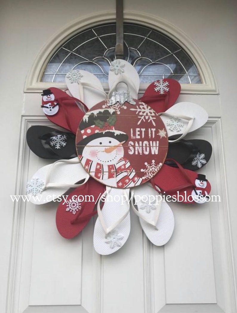 May include: A festive winter wreath made of red, white, and black flip-flops arranged in a circular pattern. A wooden ornament in the center features a snowman and the words "LET IT SNOW". Snowflake accents are visible.