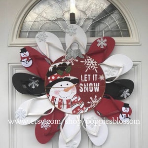 May include: A festive winter wreath made from red, white, and black flip-flops, arranged in a circular pattern. The wreath features a wooden ornament with a snowman and the words "LET IT SNOW". Decorative snowflakes and small snowman accents are also included.