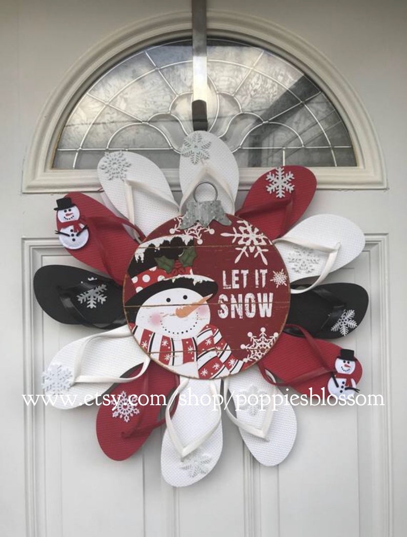 May include: A festive winter wreath made from red, white, and black flip-flops, arranged in a circular pattern. The wreath features a wooden ornament with a snowman and the words "LET IT SNOW". Decorative snowflakes and small snowman accents are also included.