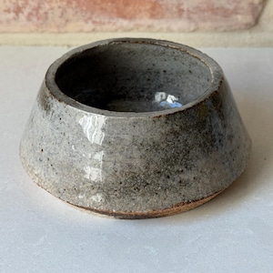 Puede incluir: Un cuenco de cerámica hecho a mano con un esmalte gris moteado. El cuenco tiene una forma ancha, ligeramente acampanada con una base redondeada y un borde marrón oscuro. El interior es de un gris más oscuro.