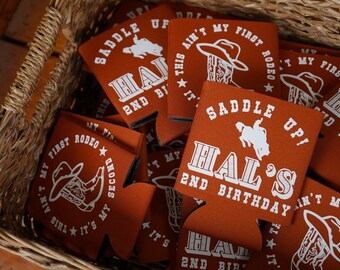 May include: A basket filled with orange drink koozies. Each koozie has white text and graphics, including cowboy hats and boots. The text reads "Hal's 2nd Birthday", "Saddle Up!", and "It's My First Rodeo".