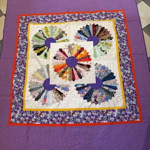 May include: A quilt featuring five fan-shaped blocks with multi-colored fabric strips and purple centers on a white background. The quilt has a yellow inner border, a floral purple and black middle border, an orange accent, and a solid purple edge.