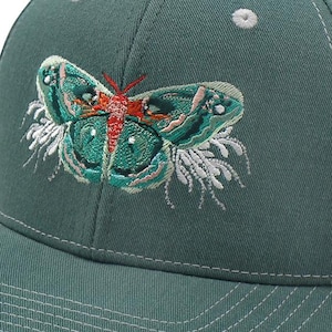 May include: A green baseball cap with a curved brim. The cap features an embroidered butterfly design in shades of green, red, and white. The cap has a button on top and white stitching details.