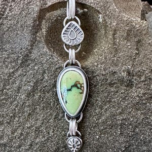 May include: A silver pendant necklace with a teardrop-shaped, light green stone. The stone is set in a silver bezel and is connected to other silver elements with decorative details. The necklace is displayed against a textured gray background.