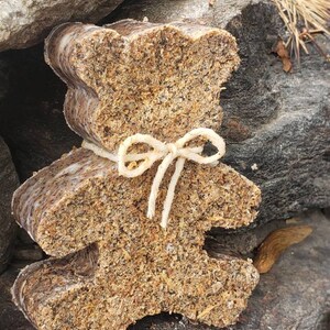 May include: A brown and beige soap bar shaped like a teddy bear with a white string tied around its neck. The soap bar is sitting on a pile of rocks.