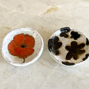 May include: Two small, round ceramic dishes. One features a vibrant orange poppy design on a speckled white background. The other has a black floral pattern on a similar speckled white surface. Both are handmade.