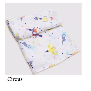 May include: White muslin baby swaddle with a colorful circus theme print. The print features a variety of circus performers, including a strongman, a clown, and a unicyclist. The swaddle is decorated with small white polka dots.