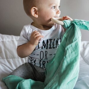 May include: A baby wearing a white onesie with the text "Eat Milk Or Try" in black. The baby is holding a light green muslin swaddle with a wooden teething ring in their mouth.