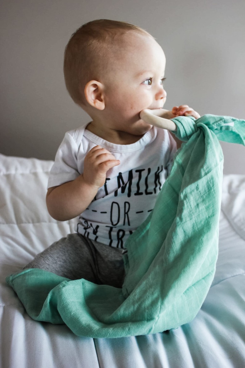 May include: A baby wearing a white onesie with the text "Eat Milk Or Try" in black. The baby is holding a light green muslin swaddle with a wooden teething ring in their mouth.
