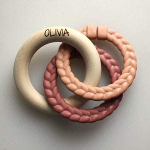 May include: A wooden teething ring with the name "Olivia" engraved on it. The ring is connected to two braided silicone rings in shades of brown and pink.