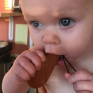 May include: A close-up of a baby with blue eyes, chewing on a wooden teething toy. The toy is brown and has a teardrop shape with a black string attached. The baby's skin is fair, and the background is blurred.