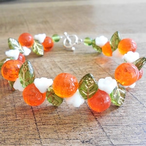 Op de afbeelding: Een armband gemaakt van oranje en groene glazen kralen, met witte kralen die op bloemen lijken. De armband heeft een zilveren sluiting.