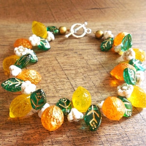 May include: A colourful beaded bracelet with yellow, orange, and green beads, and white flower-shaped beads. The bracelet has a silver-toned clasp. The beads are made of glass or resin.
