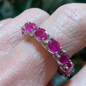 May include: A silver ring featuring a series of vibrant, round, ruby gemstones. The ring's design showcases the rich, deep red color of the rubies, creating a striking and elegant piece of jewelry. The gemstones are set closely together, forming a continuous band.