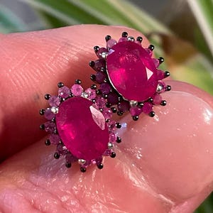 May include: A pair of ruby earrings. Each earring features a large, oval-cut ruby surrounded by smaller, pink gemstones. The earrings are set in a dark metal, creating a striking contrast with the vibrant red and pink stones. The design is elegant and eye-catching.