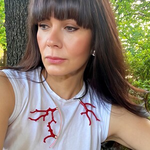 May include: A white sleeveless shirt with a coral-inspired necklace. The necklace features red, branch-like elements and a pink, curved accent. The woman in the image is outdoors, with a tree trunk and green foliage in the background.