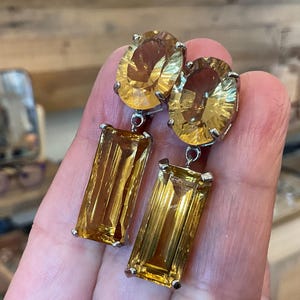 May include: A pair of dangling earrings featuring large, oval-cut, and rectangular-cut yellow gemstones. The earrings have a silver-colored metal setting. The gemstones have a warm, golden hue, and the earrings are held up by a person's hand.