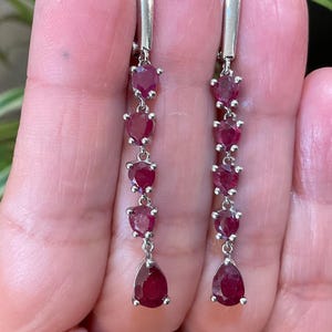 May include: A pair of dangle earrings featuring a series of heart-shaped, deep red gemstones. The earrings have a silver-colored metal setting and a teardrop-shaped ruby at the bottom. The earrings are displayed against a blurred background.