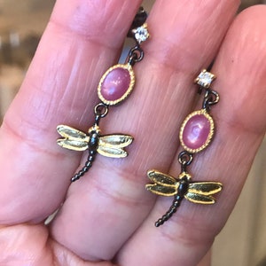 Puede incluir: Un par de pendientes colgantes con una piedra ovalada rosa y un dije de libélula dorada. Los pendientes están hechos de metal negro y dorado.