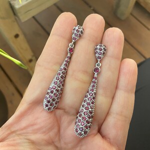 K&ouml;nnte beinhalten: Ein Paar silberfarbene Ohrh&auml;nger. Jeder Ohrring hat ein tropfenf&ouml;rmiges Design, das mit kleinen, runden, roten Edelsteinen besetzt ist. Die Ohrringe haben oben und unten einen kleinen, runden roten Edelstein. Die Ohrringe sind ca. 6 cm lang.
