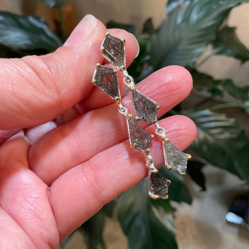 K&ouml;nnte beinhalten: Silberne Ohrringe mit einem baumelnden Design mit mehreren facettierten schwarzen Steinen mit goldenen Akzenten.