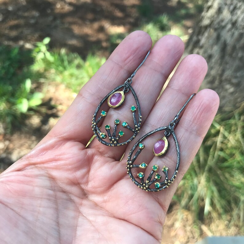 May include: A pair of dangle earrings with a teardrop-shaped design. The earrings are made of black metal with gold accents and feature pink and green gemstones.