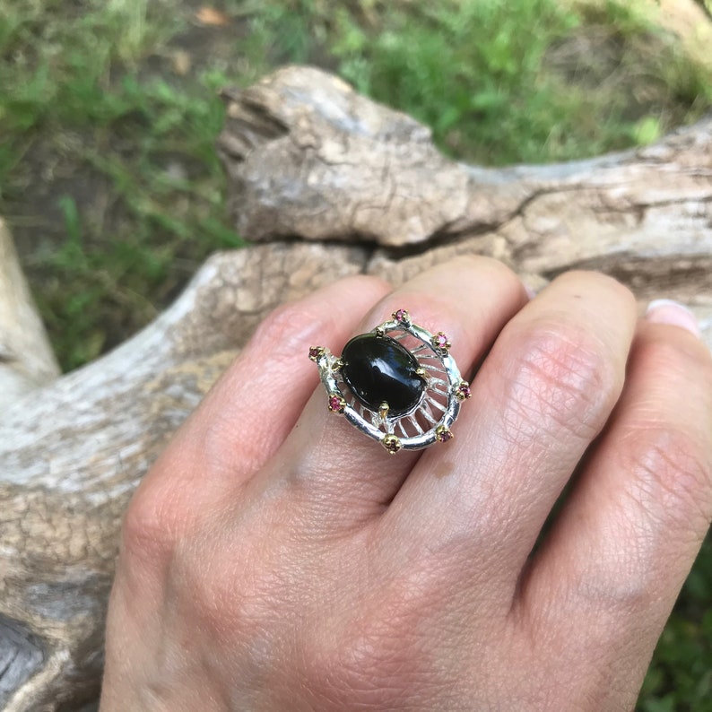 Peut inclure: Une bague en argent avec une pierre ovale noire sertie dans un motif de filigrane dor&eacute;. La bague est orn&eacute;e de petites pierres roses.