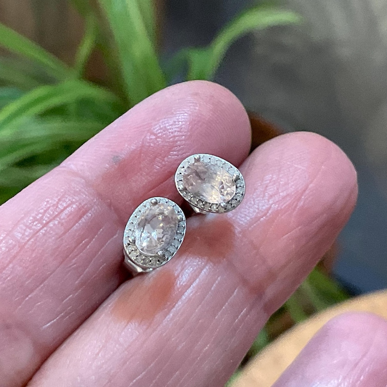 May include: A pair of oval-shaped stud earrings. Each earring features a light peach-colored gemstone surrounded by a halo of small, clear stones. The earrings are set in a silver-colored metal.