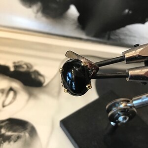 May include: A black gemstone ring with gold accents. The ring is held in a metal clamp.