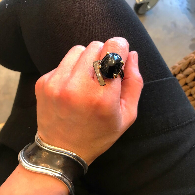 May include: A silver cuff bracelet with a wavy design. A ring with a black oval gemstone and gold accents is worn on the index finger.