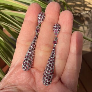 K&ouml;nnte beinhalten: Ein Paar silberfarbene Ohrh&auml;nger. Jeder Ohrring hat ein tropfenf&ouml;rmiges Design, das dicht mit kleinen, runden, tiefroten Edelsteinen bedeckt ist. Die Ohrringe haben einen kleinen, runden Edelstein oben und einen weiteren in der Mitte.