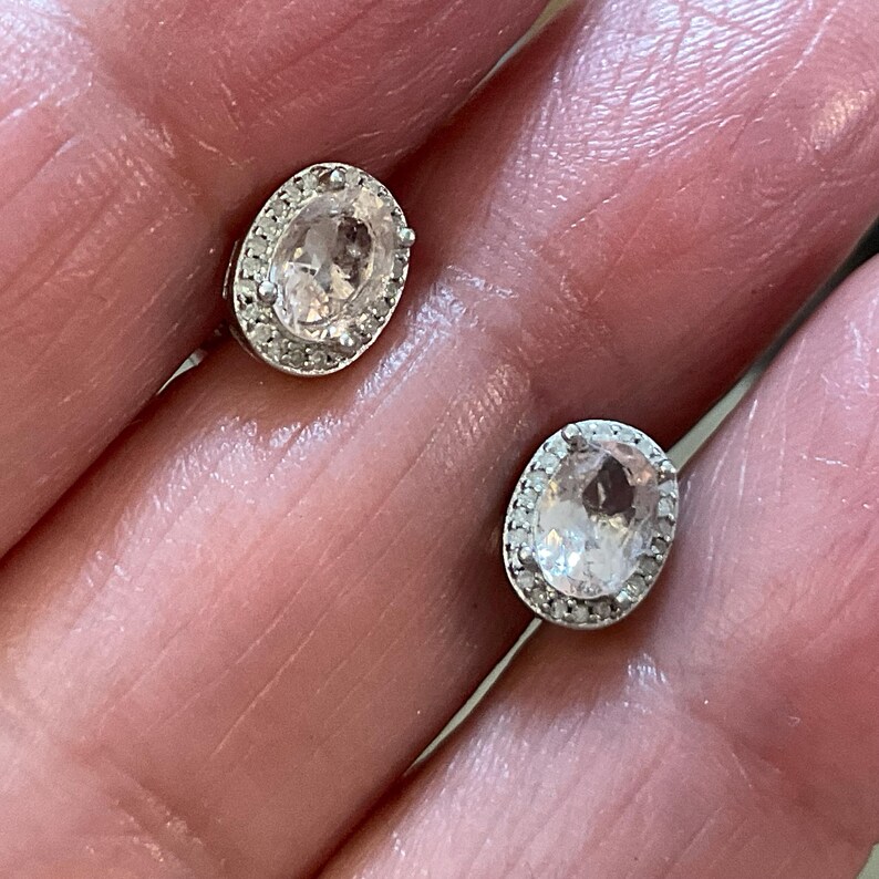 May include: A pair of oval-shaped stud earrings. Each earring features a light pink gemstone surrounded by a halo of small, clear stones. The earrings are set in a silver-colored metal.