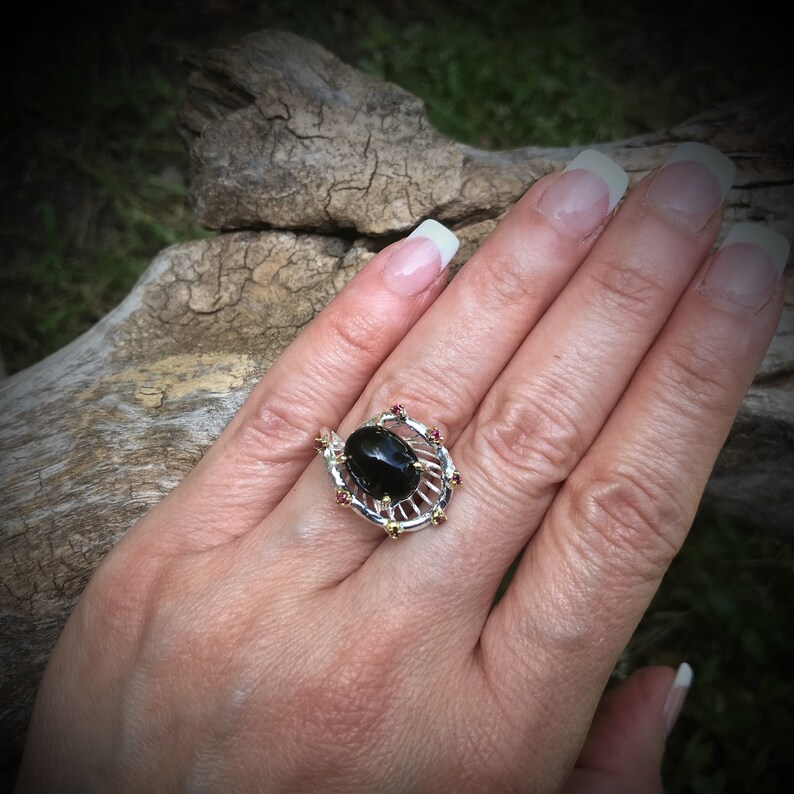 Peut inclure: Une bague en argent avec une pierre ovale noire entour&eacute;e d'un motif en filigrane dor&eacute; et de petites pierres rouges.
