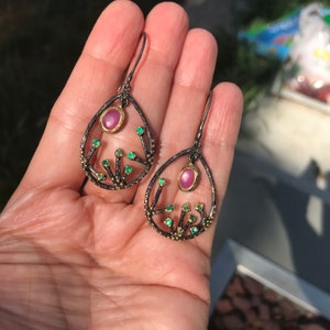 May include: A pair of dangle earrings with a teardrop-shaped frame. The frame is made of black metal with gold accents and features a pink gemstone in the center. The frame is decorated with small green gemstones.