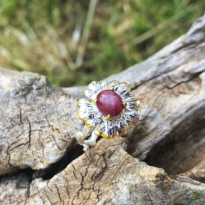 May include: A silver ring with a gold flower design and a red ruby gemstone in the center.