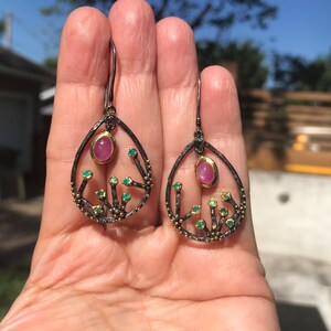 May include: A pair of dangle earrings with a black oxidized silver setting and a pink gemstone surrounded by smaller green gemstones.