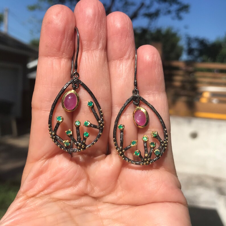 May include: A pair of dangle earrings with a black oxidized silver setting and a pink gemstone surrounded by smaller green gemstones.