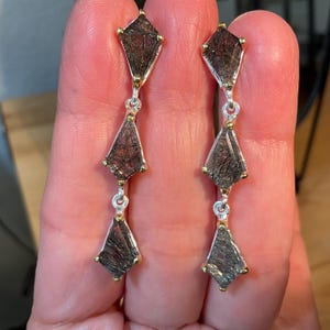 K&ouml;nnte beinhalten: Ein Paar silberne Ohrringe mit drei schwarzen und goldenen Edelsteintropfen. Jeder Tropfen ist ein facettierter, drachenf&ouml;rmiger Stein mit einem goldenen Rand.